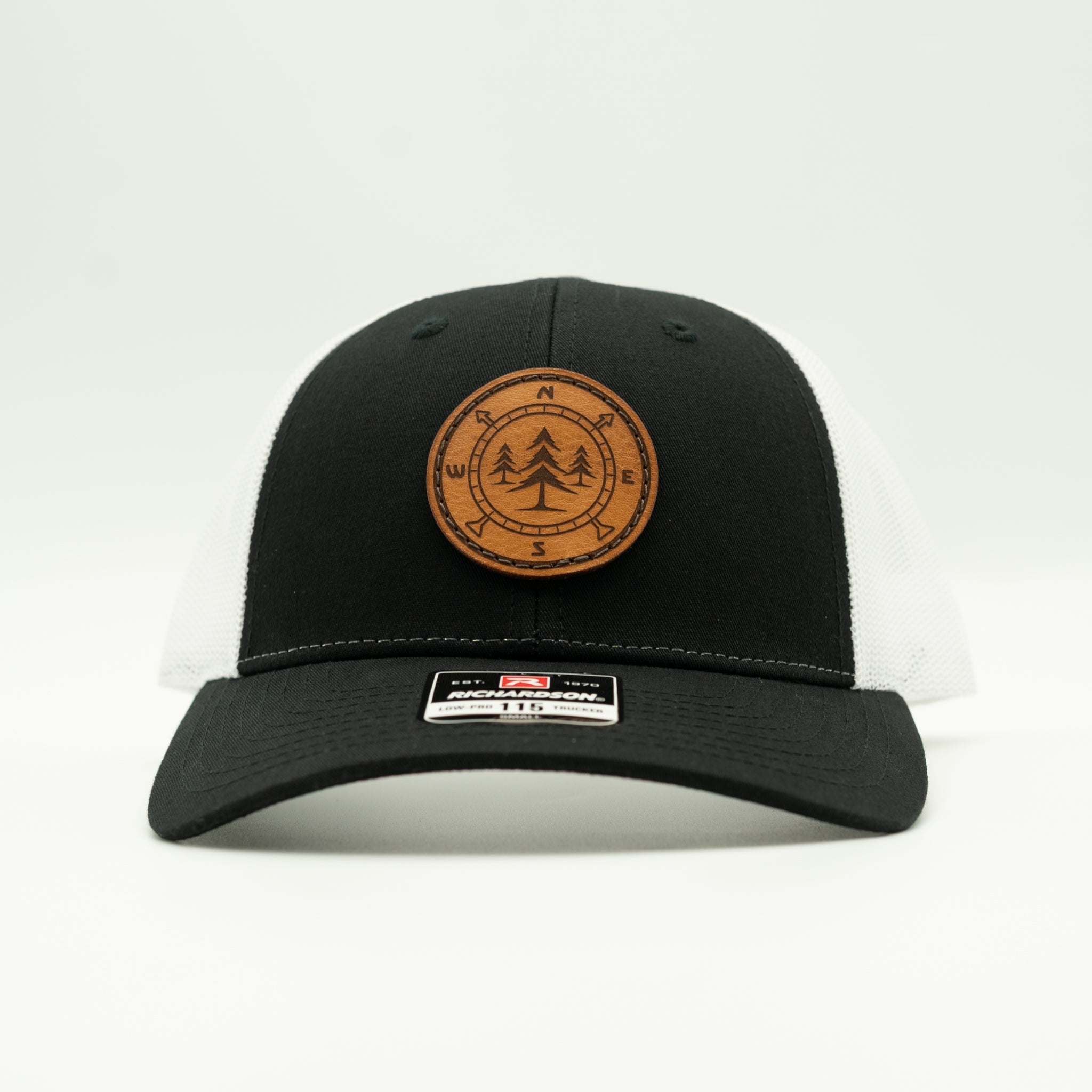 A black and white trucker hat with a brown circular leather patch on the front featuring a pine tree design, shown against a plain white background.

Color: Black/White