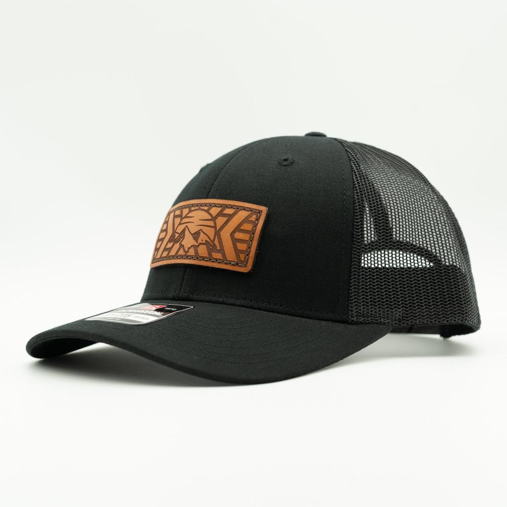 A black trucker-style cap with a brown leather patch on the front featuring a mosaic mountain design, shown against a plain white background.

Color: Solid Black