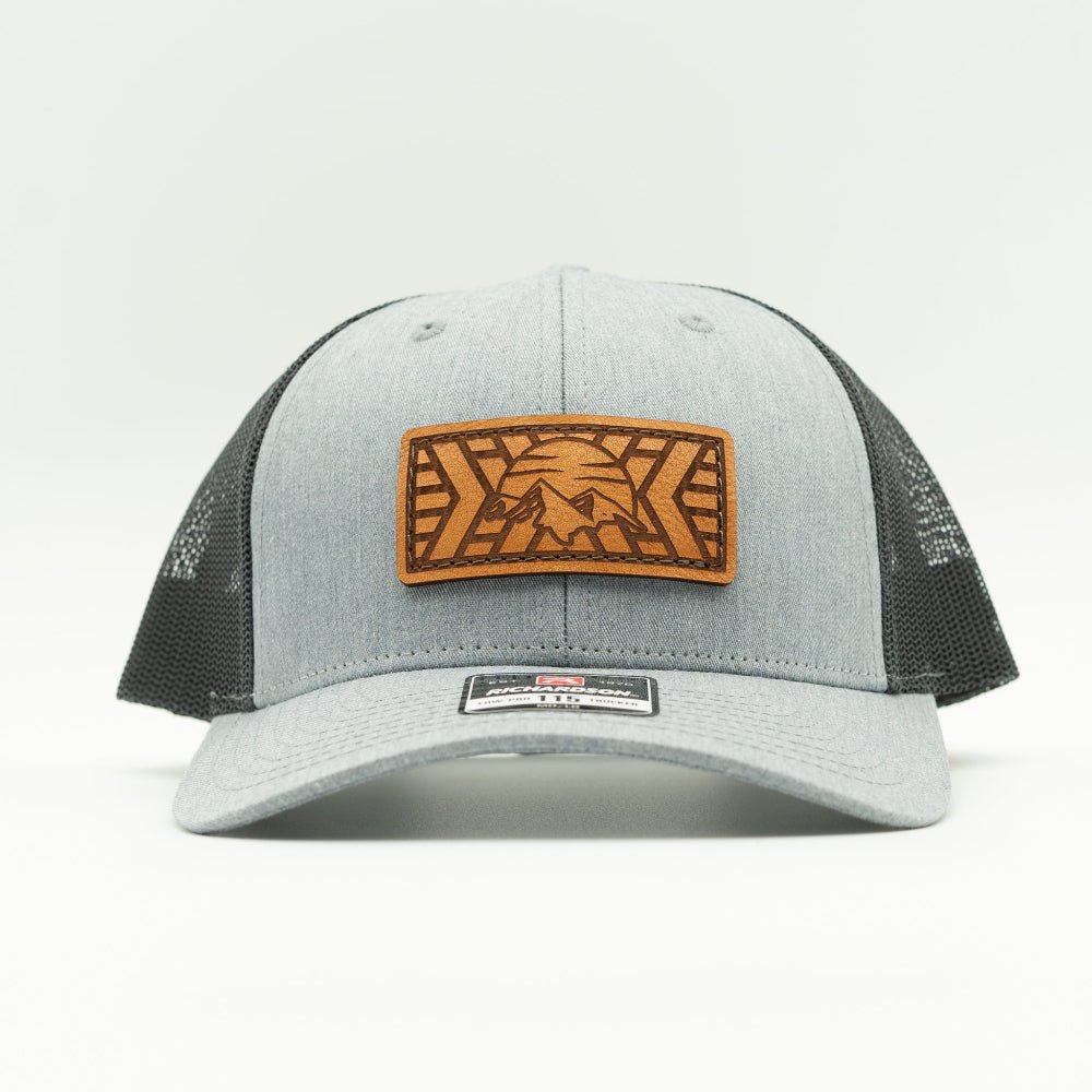 A light gray heather trucker cap with black mesh back panels and a brown leather patch on the front featuring a mountain design.

Color: Heather Grey/Charcoal