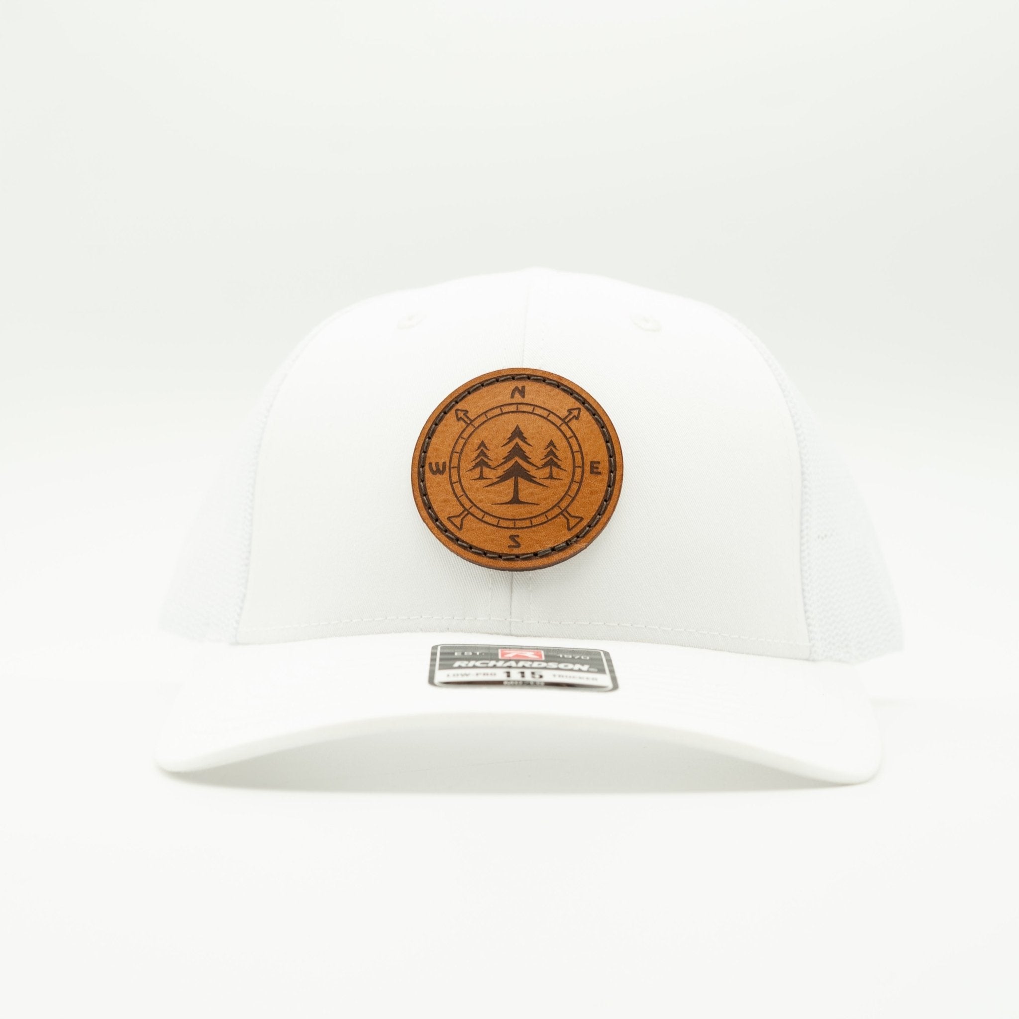 A white structured baseball cap with a circular brown leather patch on the front featuring a tree design and compass directions, photographed against a seamless white background.

Color: Solid White