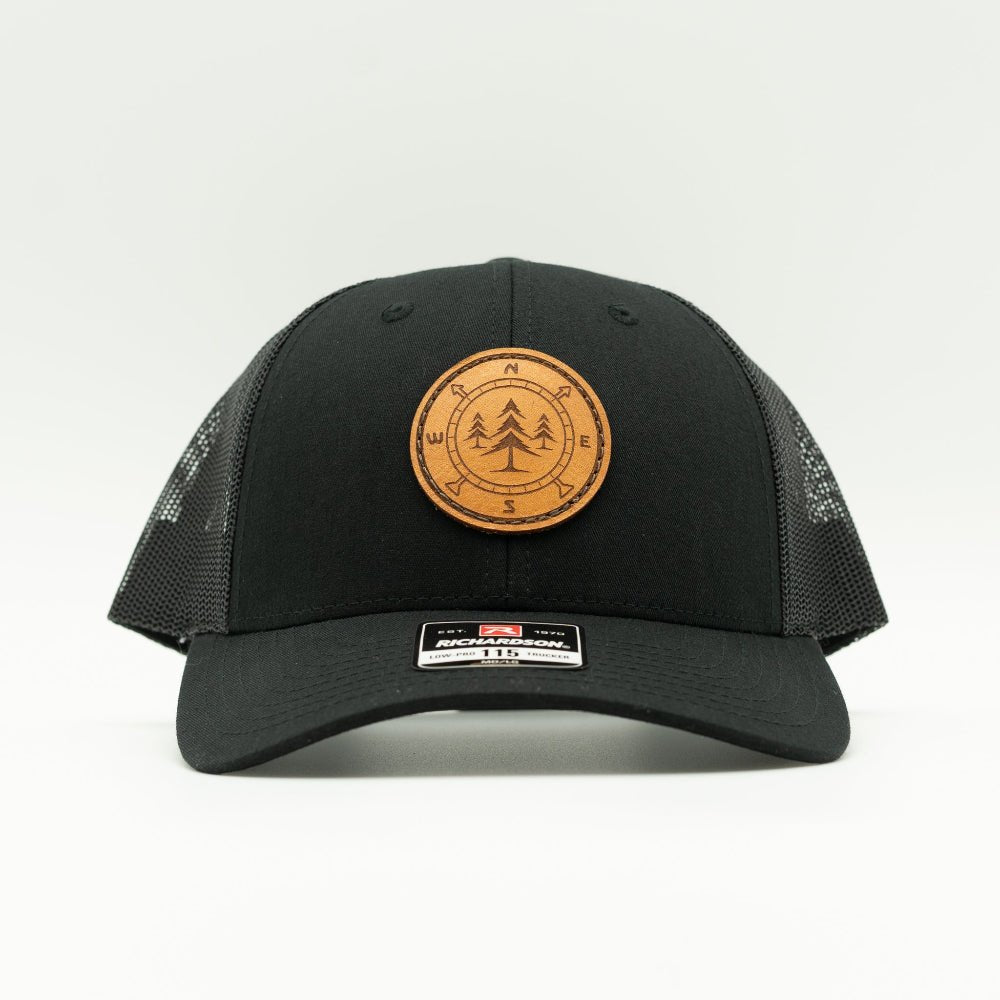 A black trucker-style cap with a circular brown leather patch featuring a tree design on the front, set against a plain light gray background.

Color: Solid Black