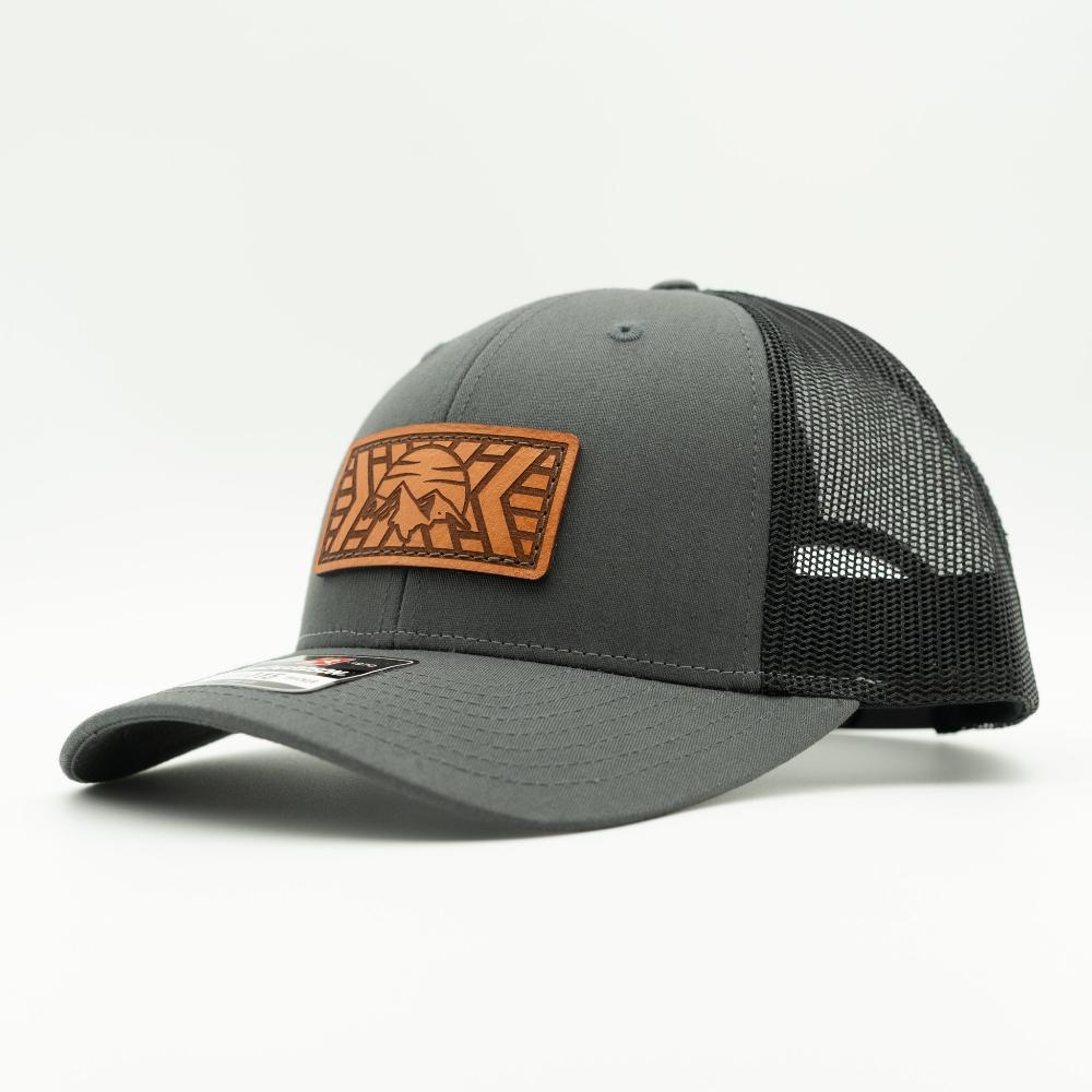 A dark gray trucker hat with black mesh back panels and a brown leather patch on the front featuring a stylized mountain and sun design.

Color: Charcoal/Black