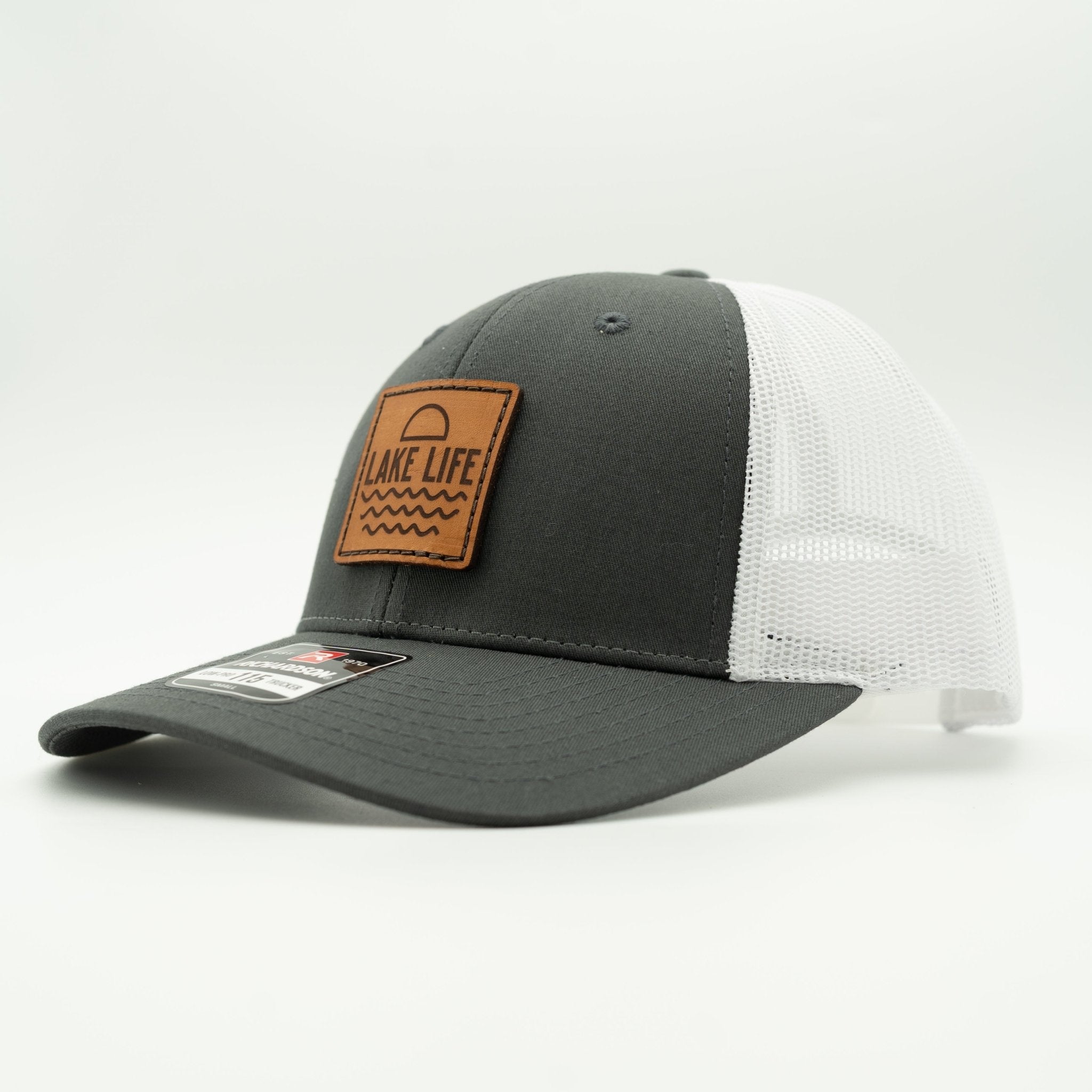 A two-tone trucker cap with a dark gray front panel and brim, white mesh back panels, and a brown leather patch on the front that reads 'LAKE LIFE!'.

Color: Charcoal/White