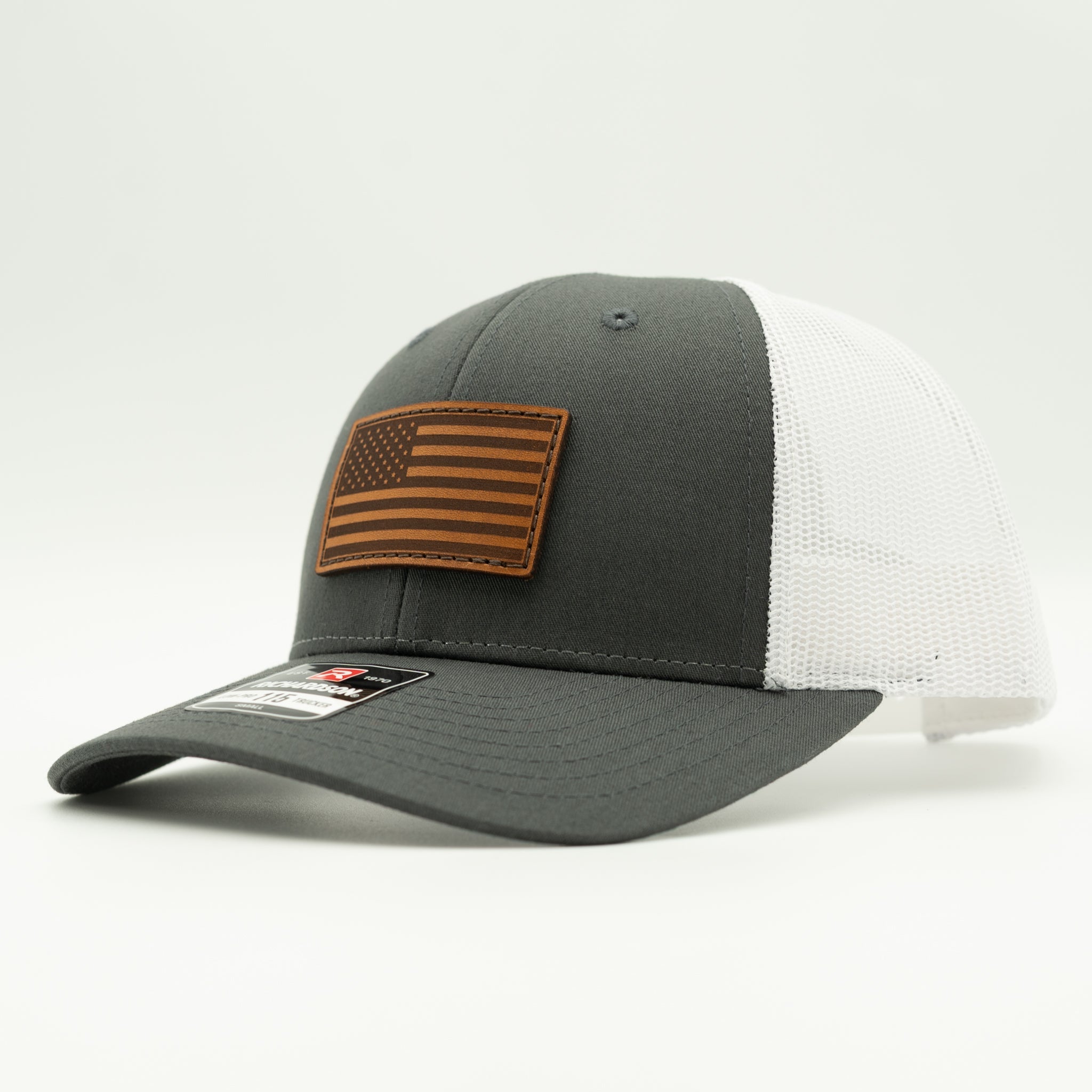 A dark gray and white trucker-style baseball cap with a brown leather American flag patch on the front, photographed on a clean white background.

Color: Charcoal/White