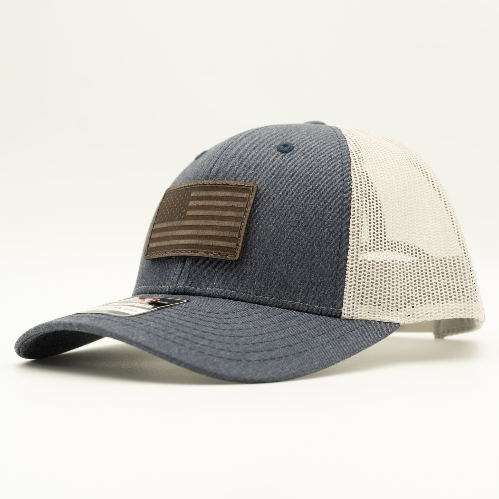 A trucker-style baseball cap with a dark heather gray front panel and brim, off-white mesh back panels, and a brown American flag patch on the front.

Color: Heather Navy/Grey