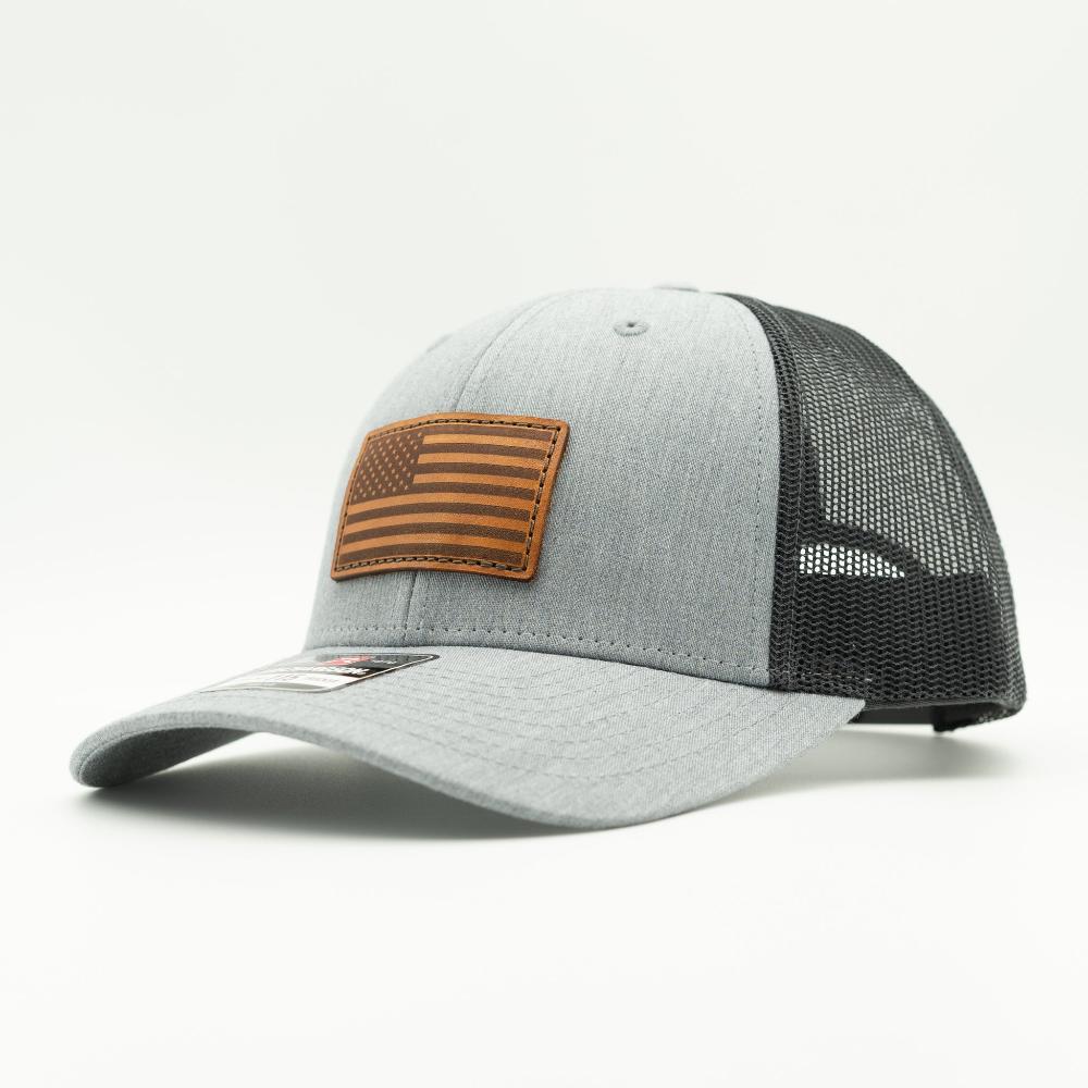 A light gray heather trucker cap with a black mesh back and a brown leather American flag patch on the front, shown on a clean white background.

Color: Heather Grey/Charcoal