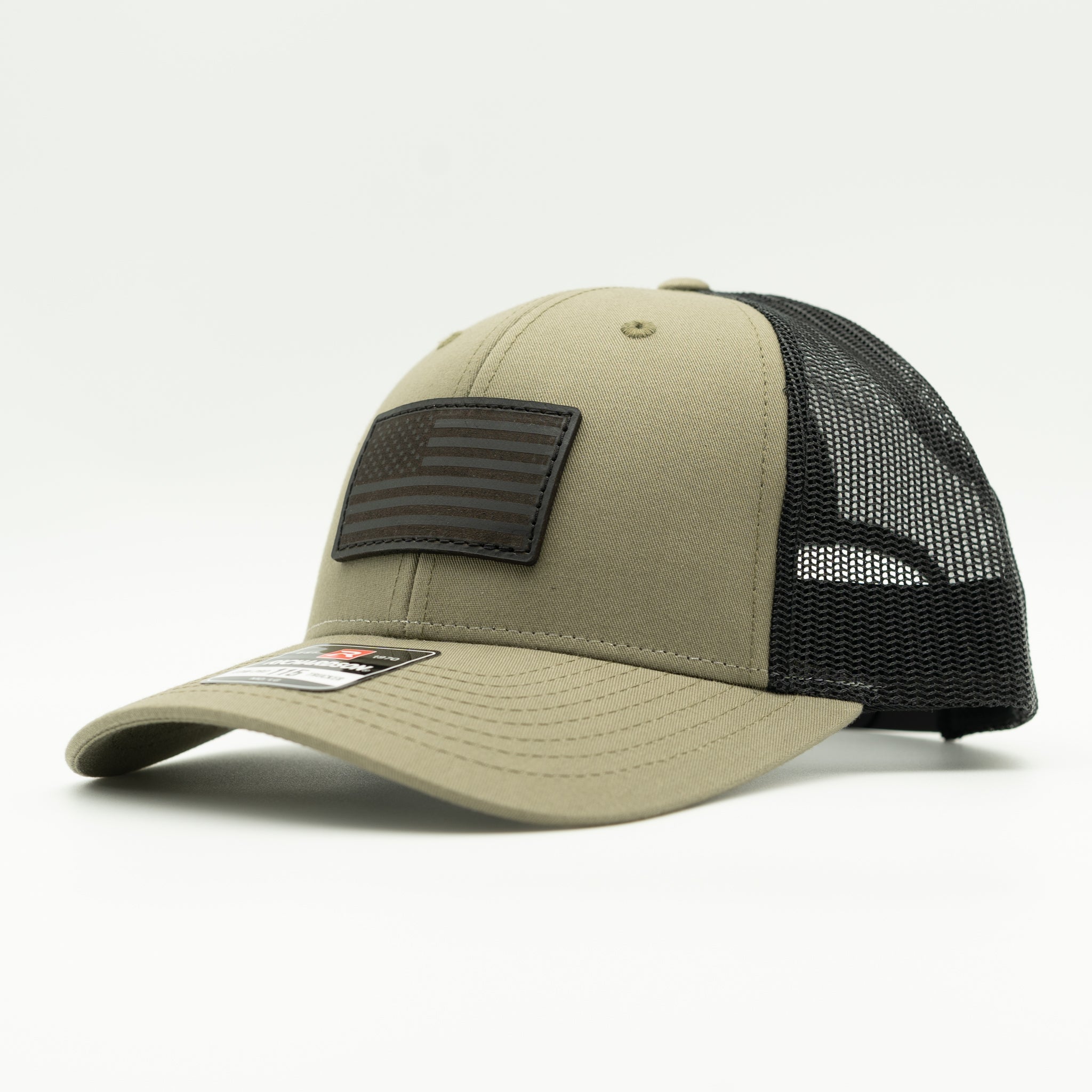 A trucker-style baseball cap with a tan/khaki front panel and brim, black mesh back, and a black rectangular American flag patch on the front, photographed on a clean white studio background.

Color: Loden/Black