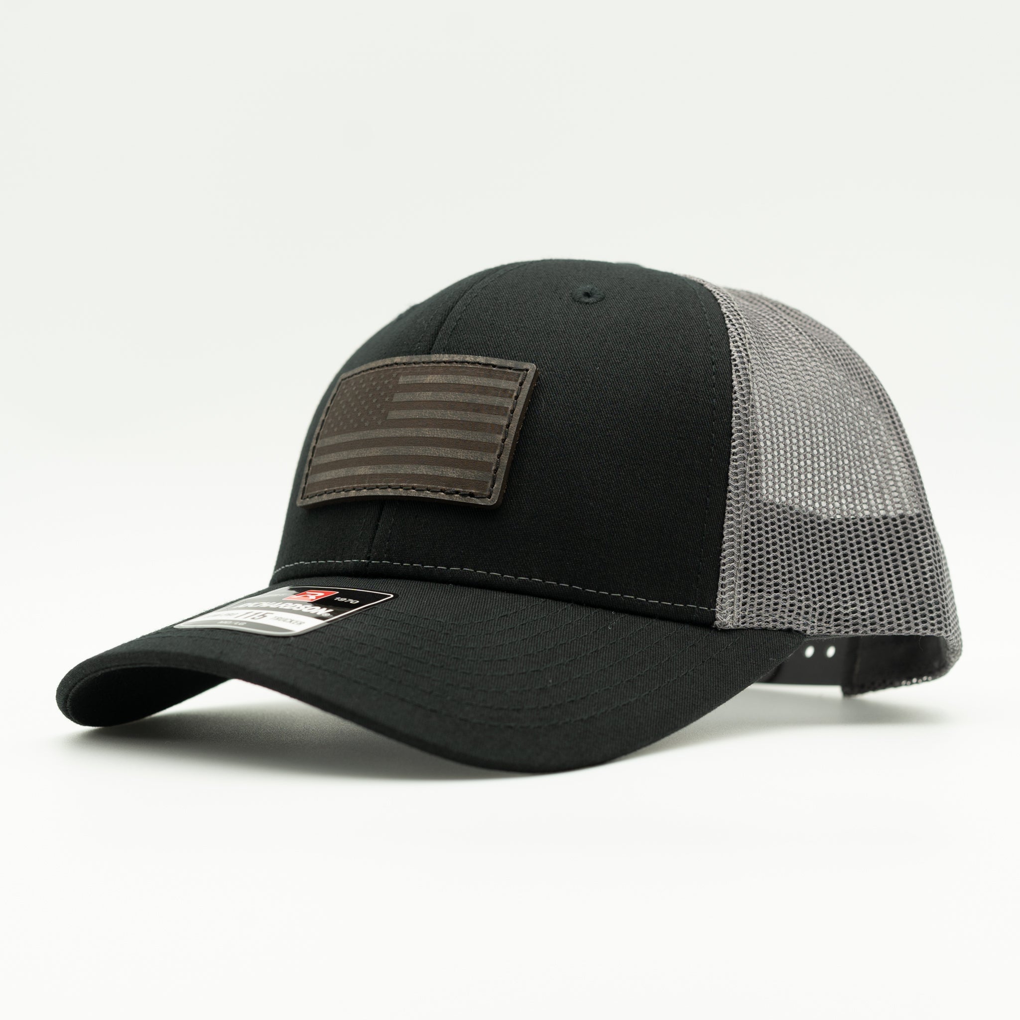A black trucker-style baseball cap with a curved brim, featuring a dark brown American flag patch on the front and a gray mesh back, shown on a light gray background.

Color: Black/Charcoal