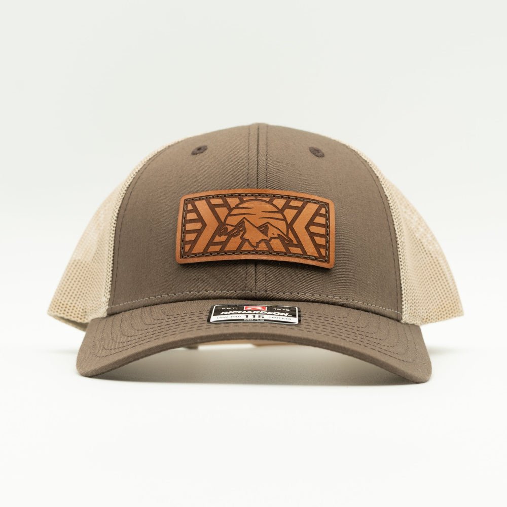 A brown and beige trucker hat with a brown leather patch on the front featuring mosaic mountain engraved patch, photographed against a clean white background.

Color: Brown/Khaki