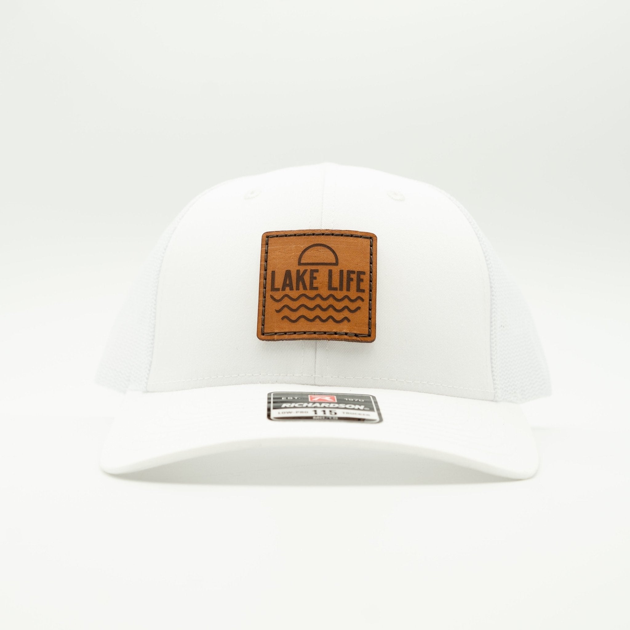 A white structured baseball cap with a brown leather patch on the front that reads 'LAKE LIFE' with a sun and wave design. A metal size tag is visible on the brim.

Color: Solid White
