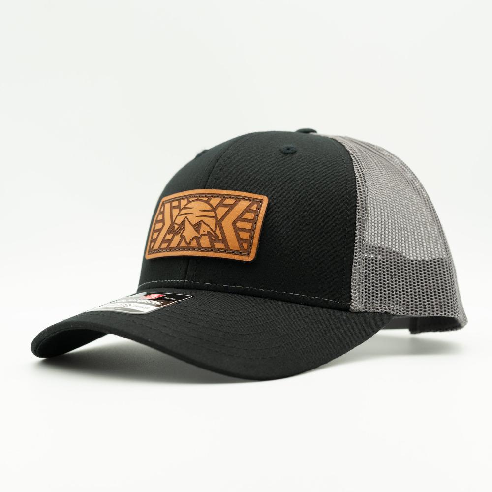 A black trucker-style cap with a tan leather patch on the front featuring a stylized mountain and sun design, and a gray mesh back, shown on a white background.

Color: Black/Charcoal
