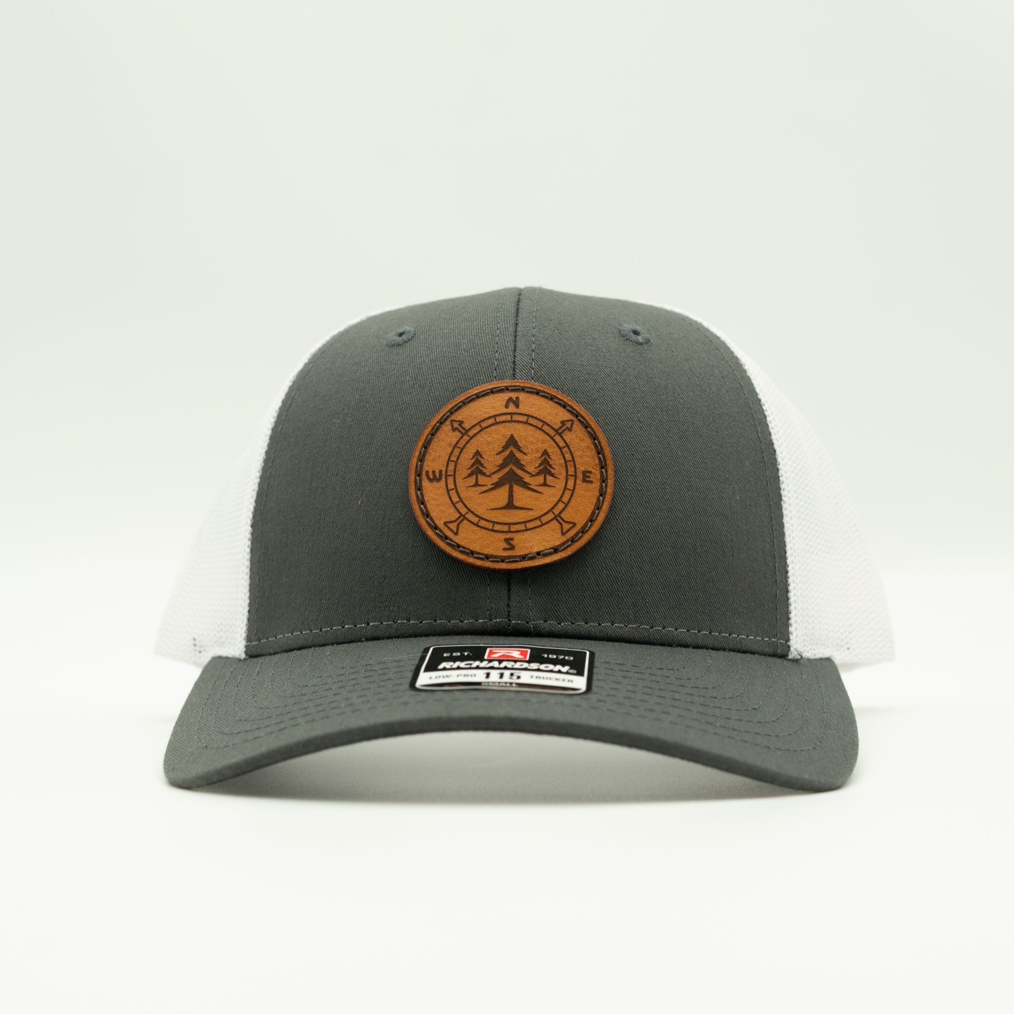 A dark gray and white trucker hat with a brown circular wooden patch on the front featuring a tree design and compass directions. 

Color: Charcoal/White