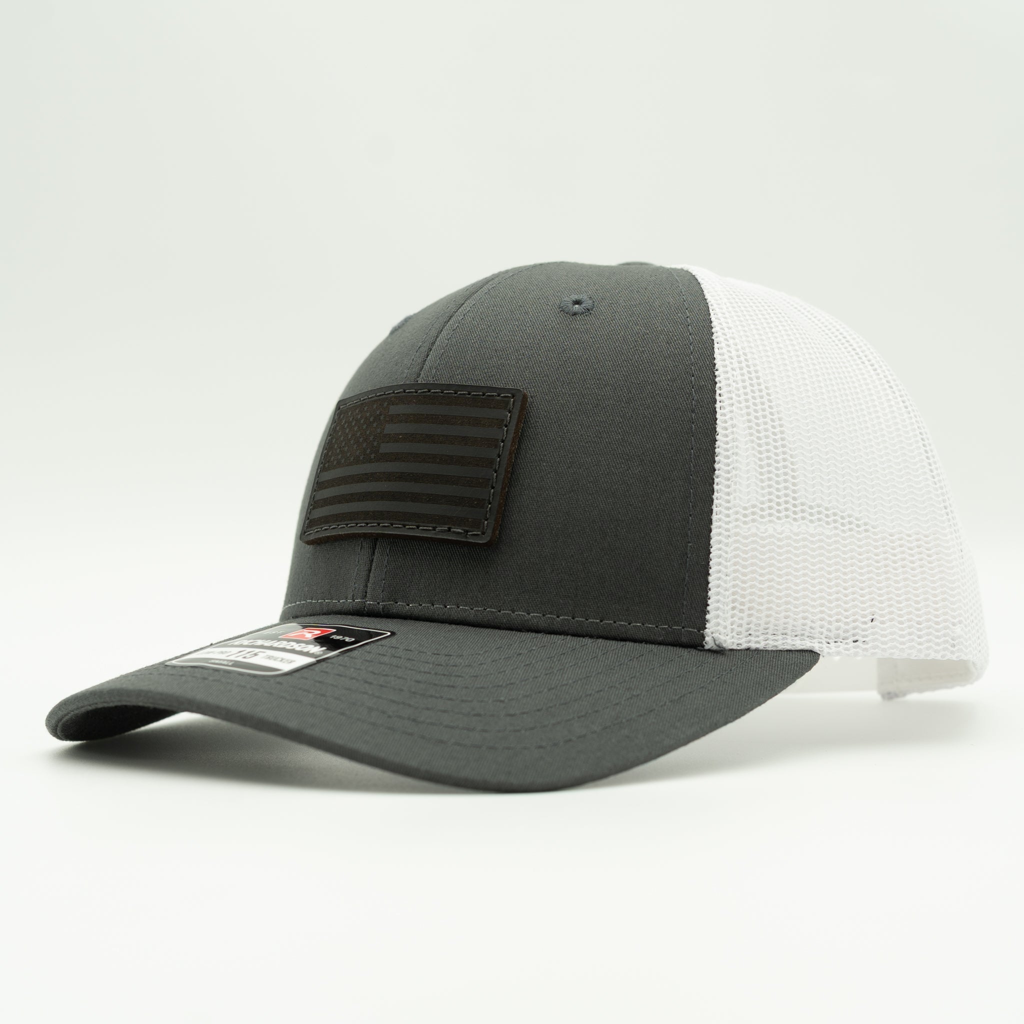A two-tone trucker cap with a dark gray front panel and brim, white mesh back panels, and a dark gray rectangular American flag patch on the front.

Color: Charcoal/White