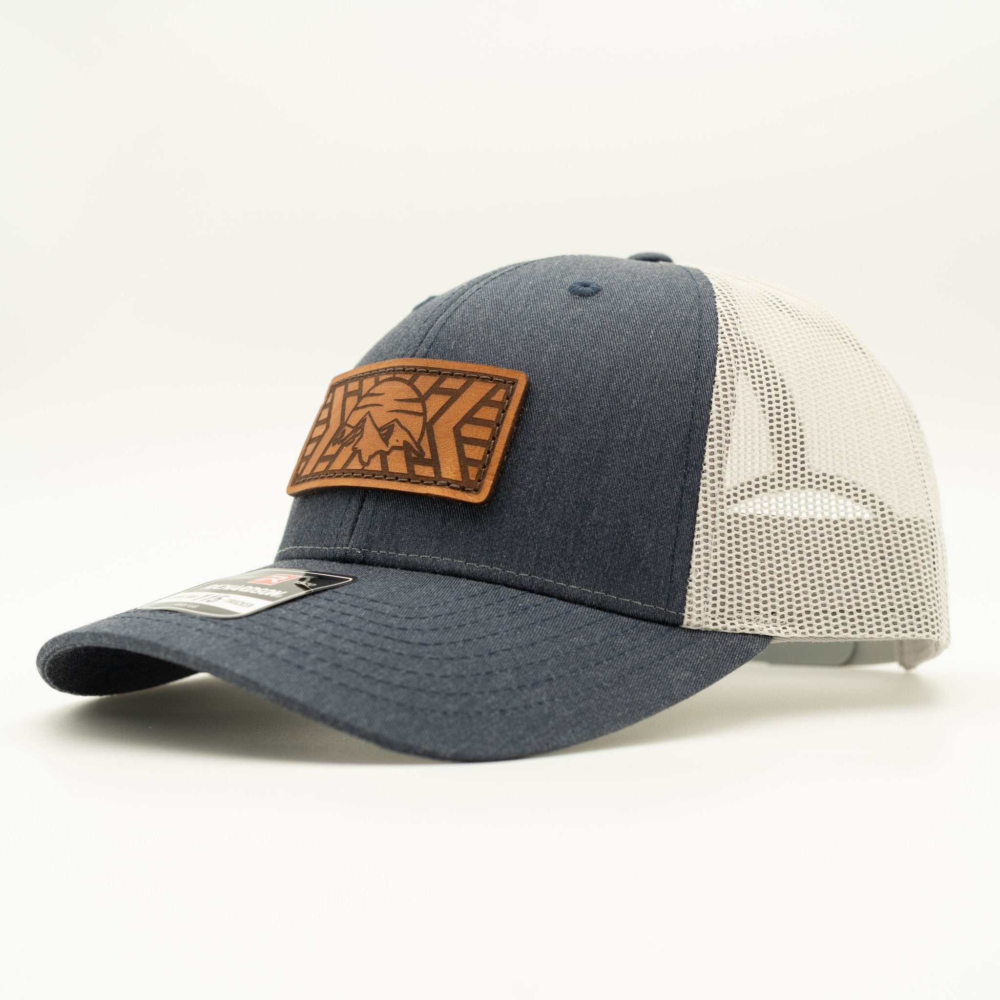 A trucker-style baseball cap with a dark heather gray front panel and brim, off-white mesh back panels, and a brown leather patch on the front featuring a stylized graphic.

Color: Heather Navy/Grey