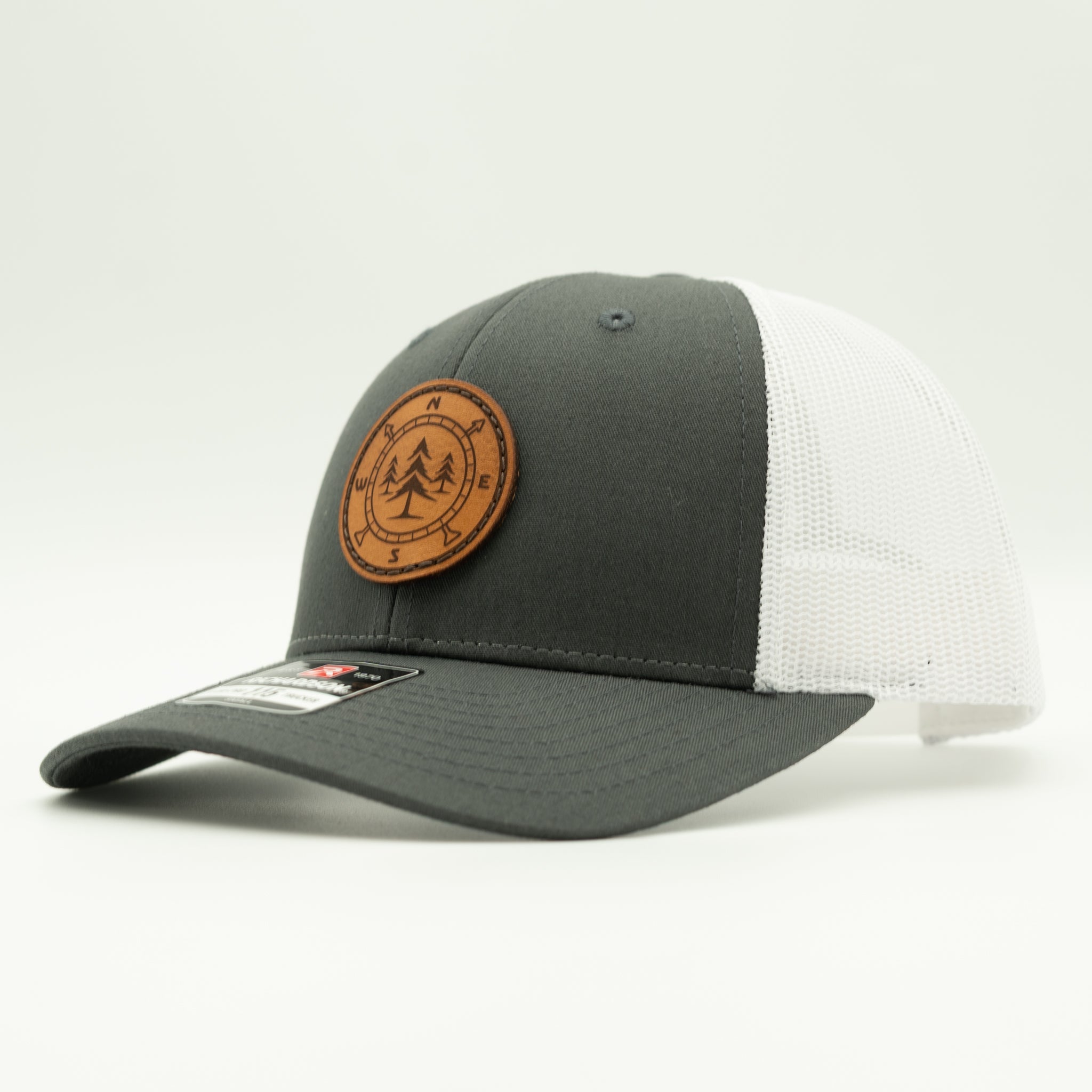 A dark gray and white trucker hat with a brown circular leather patch on the front, photographed on a clean white background.

Color: Charcoal/White