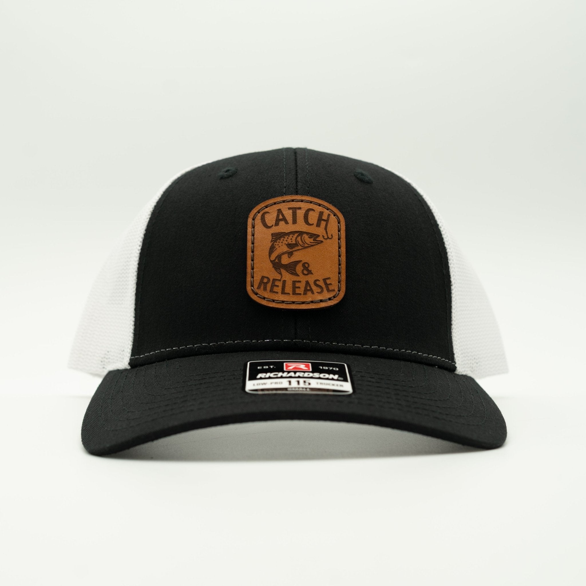 A black and white trucker hat with a brown leather patch on the front that reads 'CATCH RELEASE' with a fish graphic. The hat has a black structured front panel, black brim, and white mesh back panels, photographed on a light gray background.

Color: Black/White