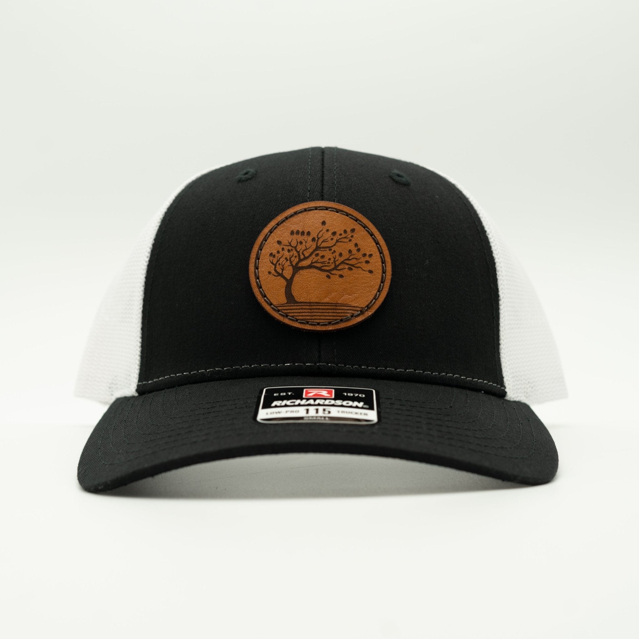 A black and white mesh back trucker hat with a brown circular leather patch on the front featuring an engraved tree design. A silver and black Richardson brand sticker is visible on the brim.

Color: Black/White