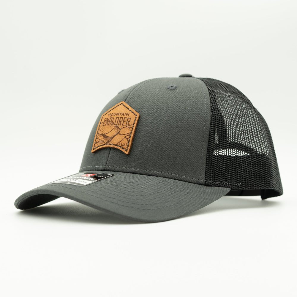 Side angle of Mountain Explorer leather patch hat on charcoal and black Richardson 115 low profile trucker, showing structured crown, mesh back, and curved bill.