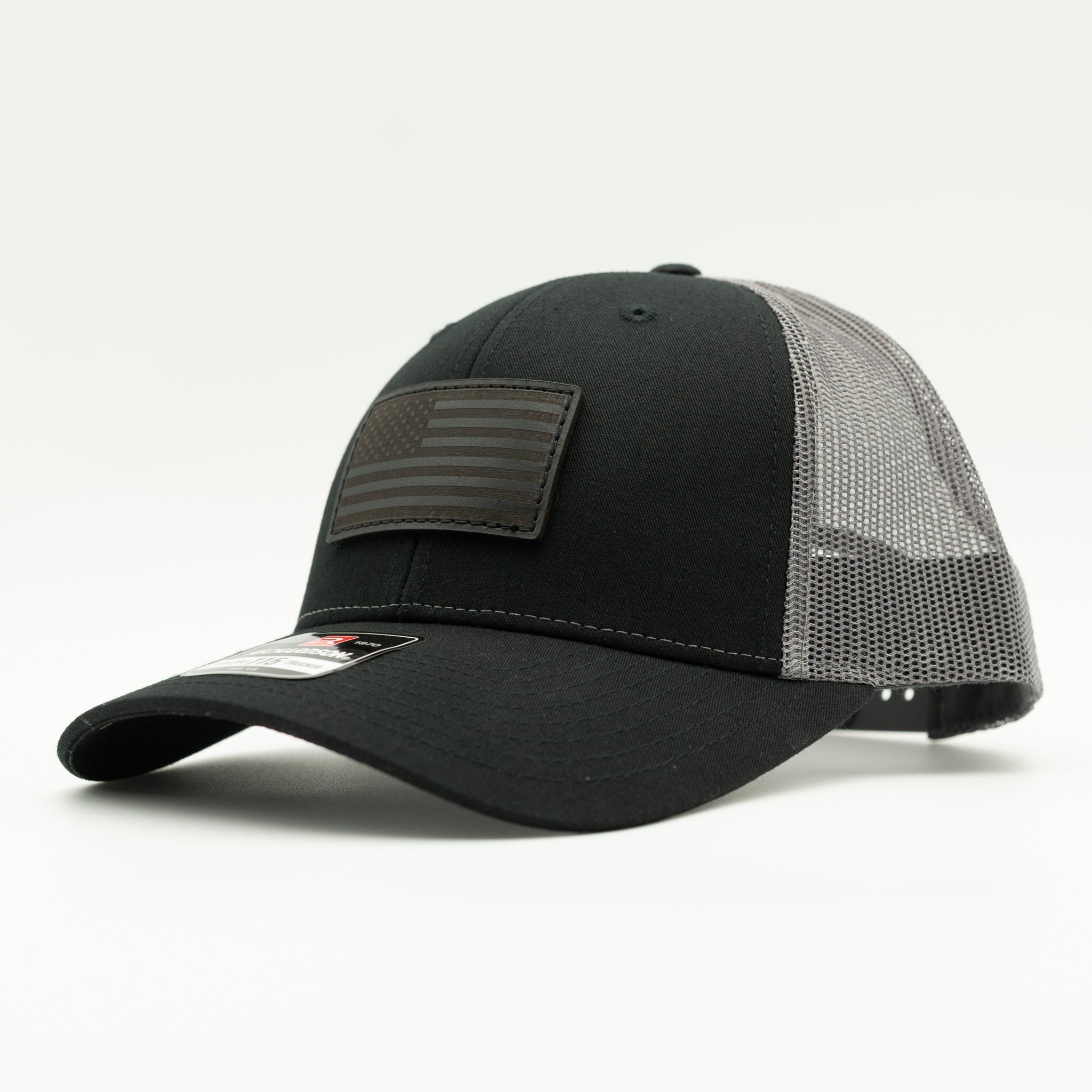 A black trucker-style baseball cap with a black mesh back and a rectangular patch featuring a faded American flag on the front, photographed on a light gray background.

Color: Black/Charcoal