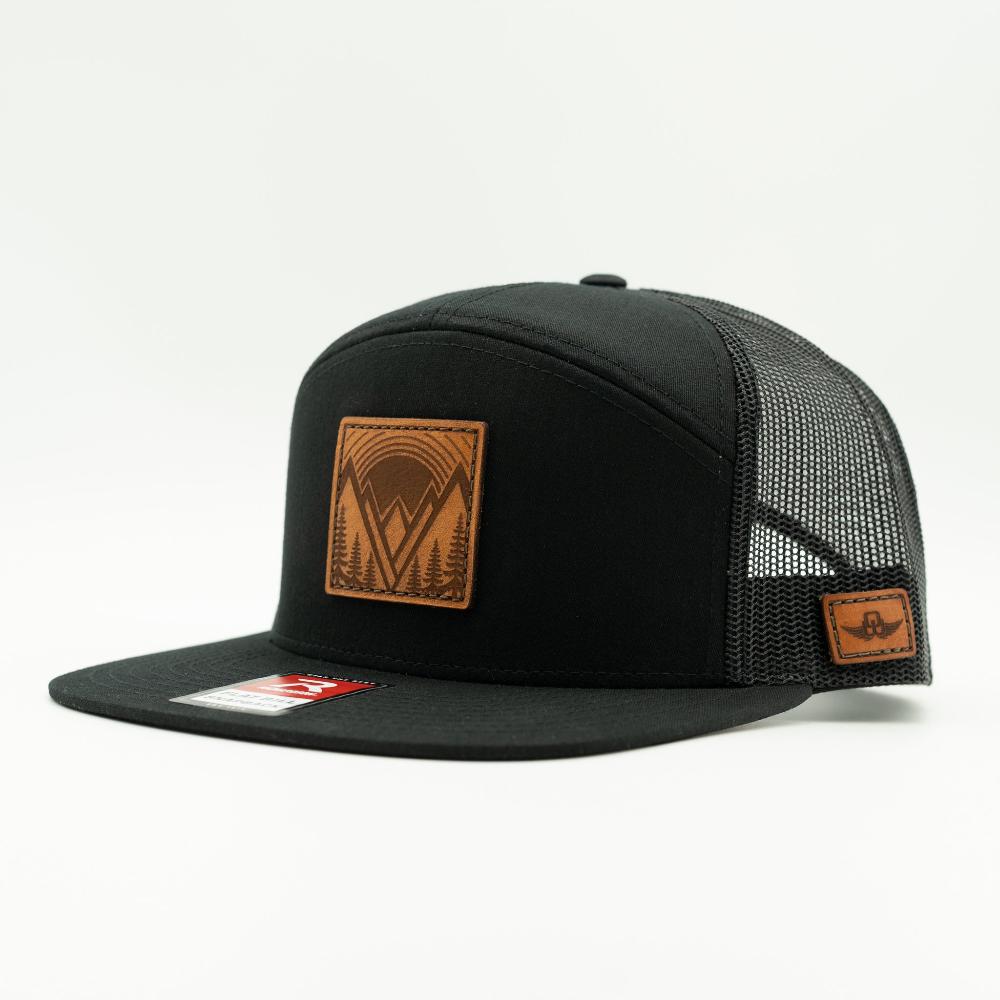 Side view of Mountain East leather patch hat on solid black Richardson 168 flat bill trucker, showing seven panel crown, mesh back, and sewn leather mountain patch.