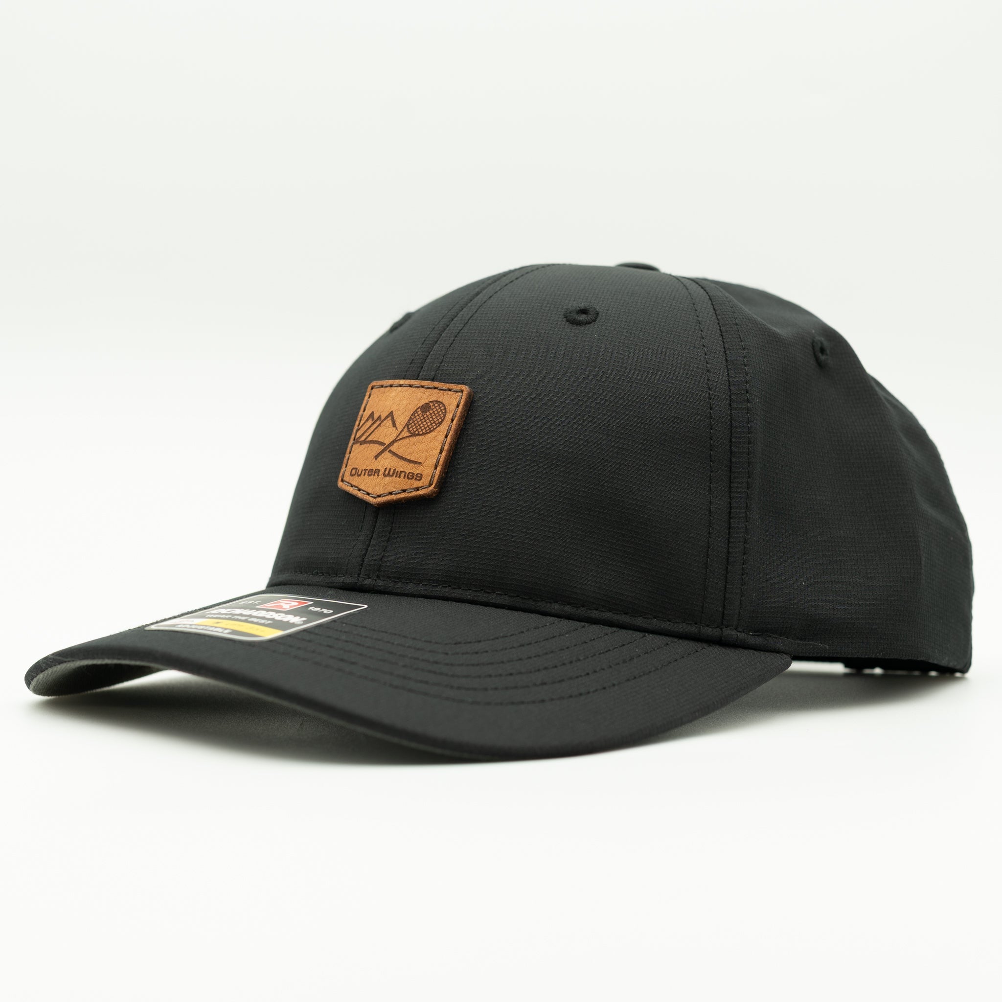 A black baseball cap with a brown leather patch on the front featuring a mountain and tennis racket graphic, photographed on a clean white background.

Color: Black