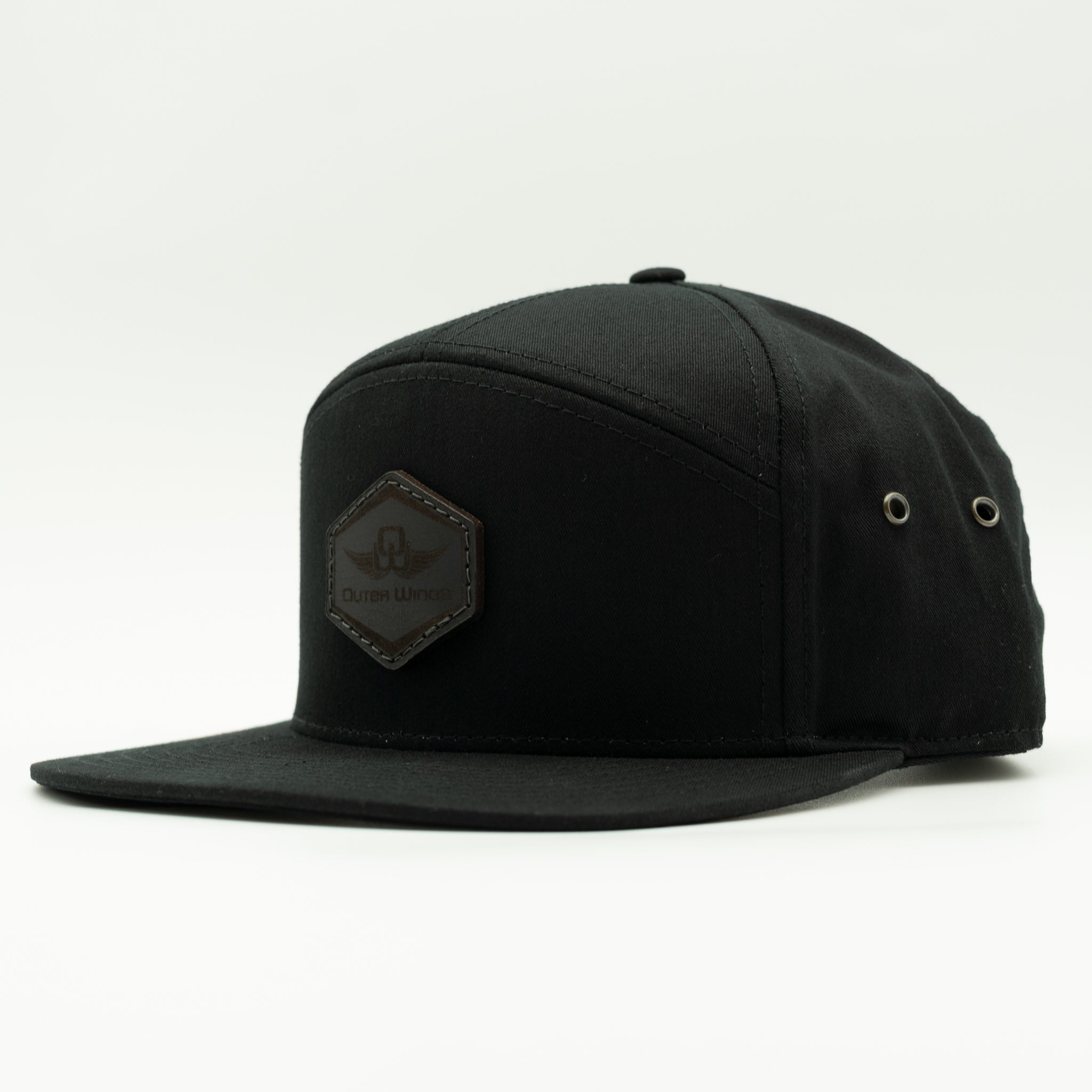 A black flat-brim snapback cap with a Black hexagonal patch on the front featuring a stylized logo and the text 'Outer Wings'. The cap is photographed on a clean, light gray studio background.

Color: Black