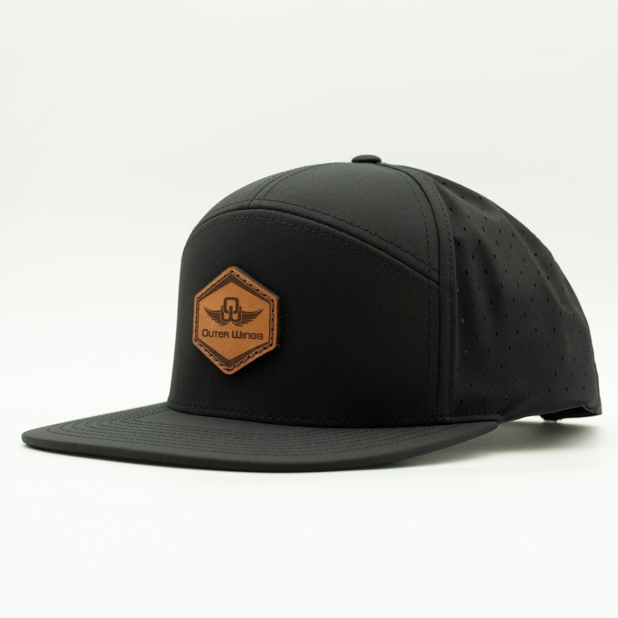A black flat-brim cap with a hexagonal brown leather patch on the front featuring the text 'Otter Wansell' and a winged logo, photographed on a clean white background.

Color: Black