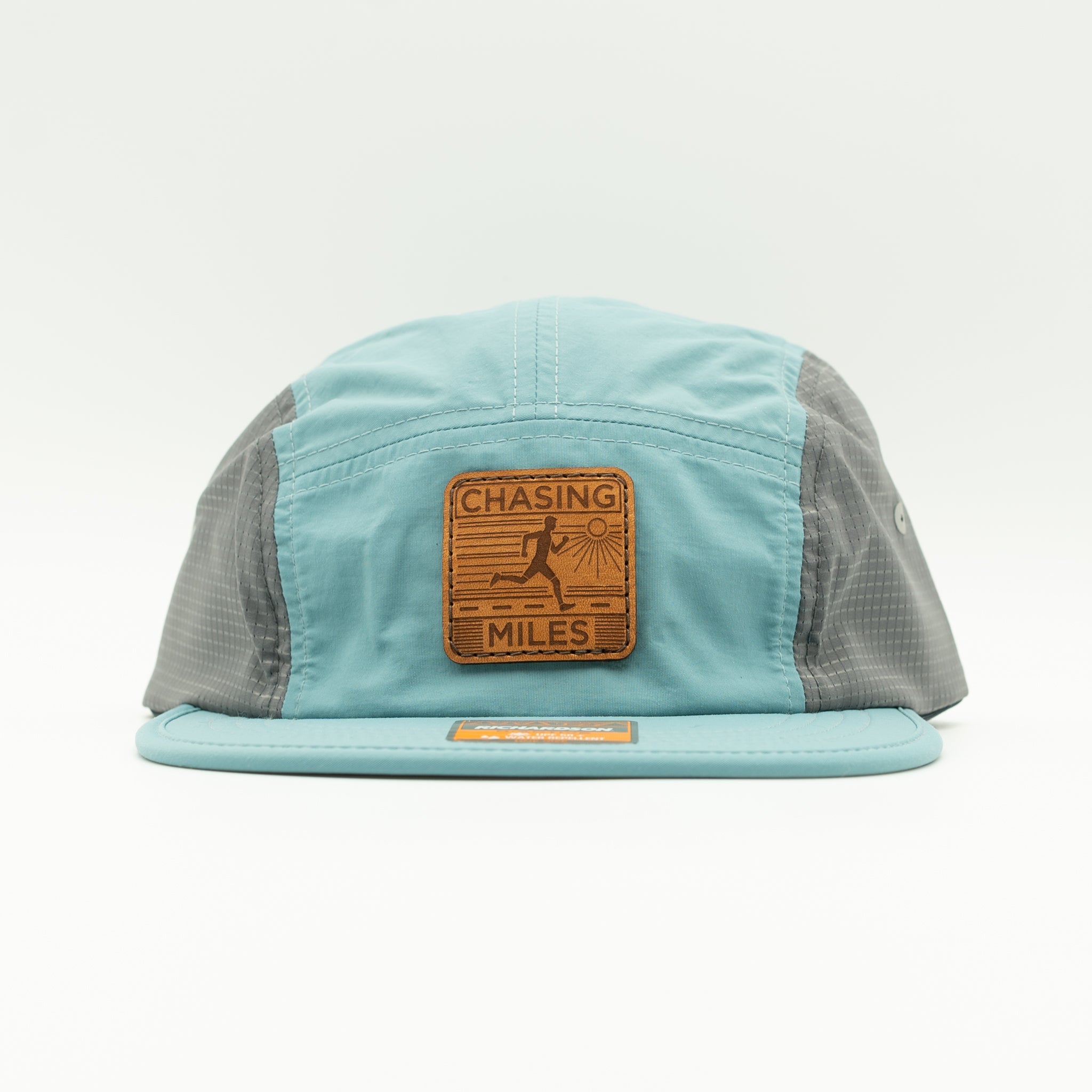 A light blue and gray 5-panel cap with a brown leather patch on the front that reads 'CHASING MILES'.

Color: Smoke Blue/Charcoal