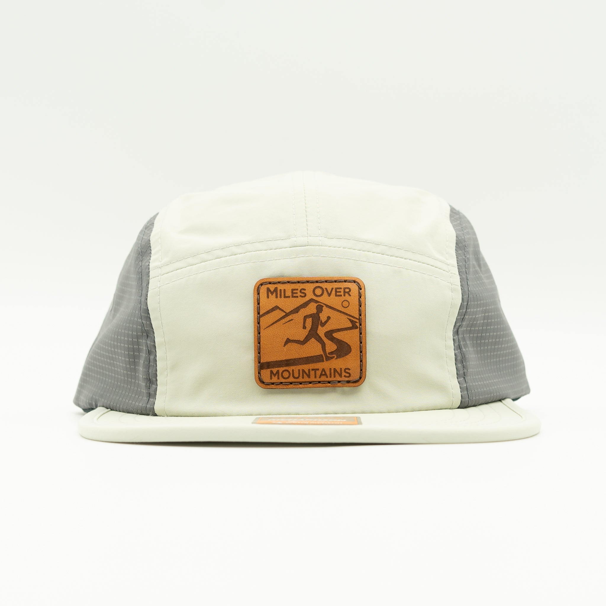A 5-panel trucker cap with an off-white front panel and brim, gray mesh side panels, and a brown leather patch on the front that reads 'MILES OVER MOUNTAINS' with a mountain and skier graphic.

Color: Light Jade/Charcoal